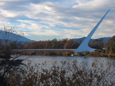 [A white bridge with a tall mast at one end from which cables are strung to support the bridge spanning the river. The sky has quite a few clouds and it's near dusk, so there is not a lot of light on the bridge or the water.]